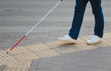 Persona con discapacidad visual utilizando un bastón blanco y un pavimento podotáctil para guiarse en una ciudad