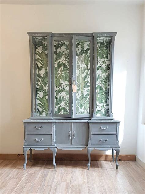 Foto de la misma vitrina de madera oscura restaurada, pintada de un tono neutro con papel pintado de motivos vegetales en el interior y una base de madera moderna